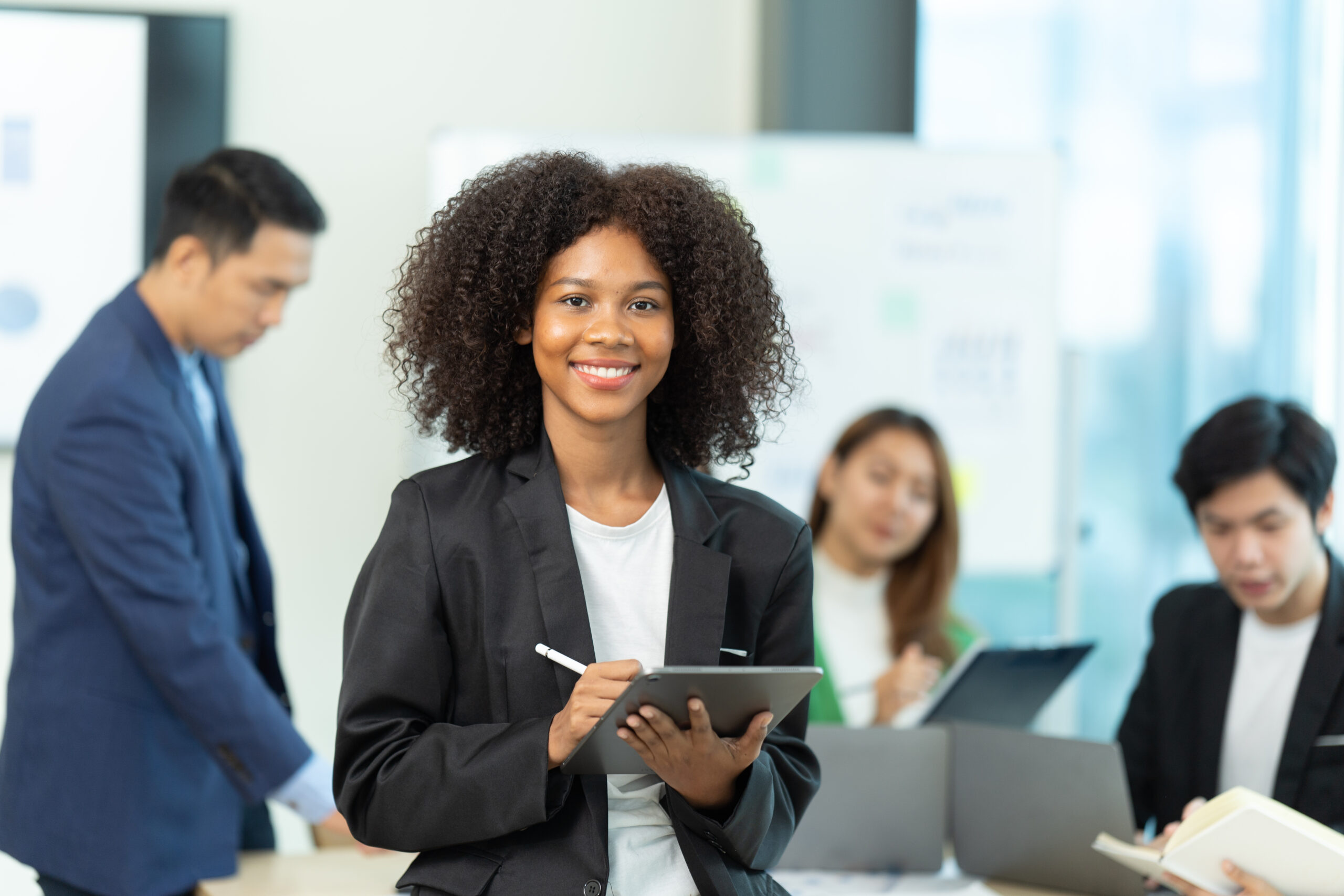 A young sales intern working on a tablet with her colleagues brainstorming in the background.